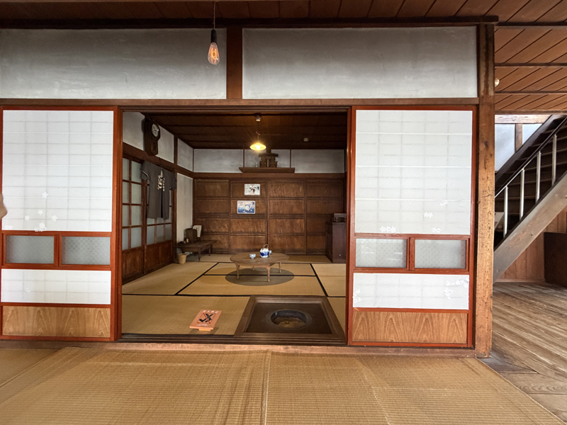 旧来正旅館の1階茶の間。手前に囲炉裏、中央にちゃぶ台と急須、壁には柱時計や半纏、上部に神棚が祀られた畳敷きの空間