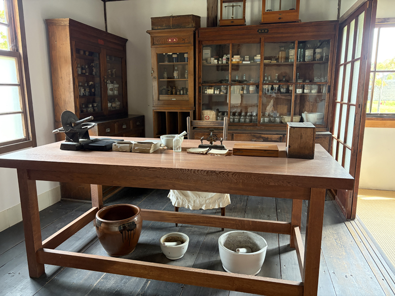 旧近藤医院の薬局（調剤室）の様子。中央の大きな木製作業台の上には、手動で錠剤を作るための黒い打錠機（車輪のようなハンドル付き）、乳鉢、薬包紙が置かれている。奥の棚には大小様々な薬瓶がずらりと並び、当時の調剤作業が再現されている