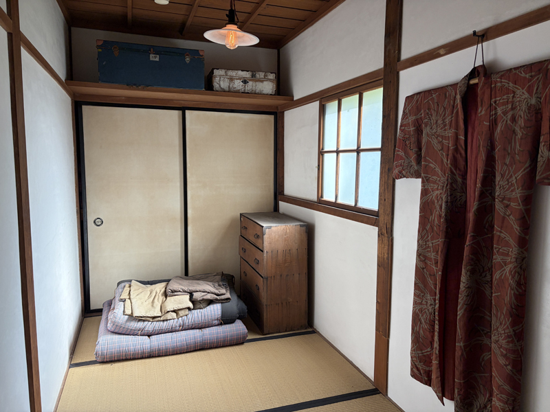 旧来正旅館の2階客室。畳の上にたたまれた布団、木製箪笥、壁に掛けられた柄物の着物、天袋に置かれた革製トランクと行李
